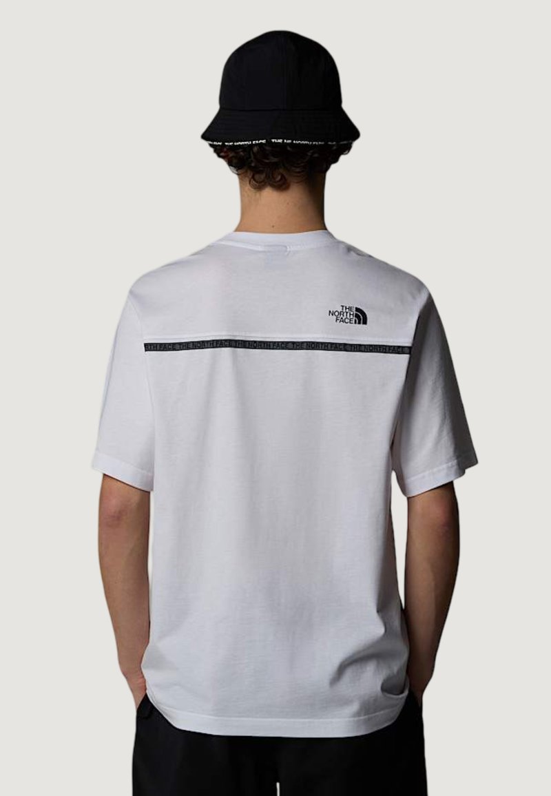 Persona con una camiseta blanca de The North Face con logo negro y franja en la parte superior de la espalda, y un sombrero tipo bucket negro, de espaldas.