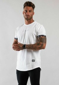 Vit t-shirt med korta ärmar som har en grafisk utskrift på ärmsluten, rak fåll och en logotagg längst ner till vänster.