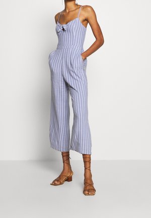 Combinaison rayée bleu clair et blanc avec un détail en nœud, des bretelles réglables, des jambes larges et des poches latérales. Portée avec des sandales à lanières camel.