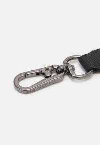 Cinghia in tessuto nero testurizzato con un gancio in metallo scuro a moschettone con la scritta "KARL LAGERFELD" incisa, collegato a un anello rotondo.