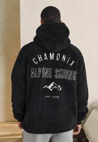 Juodas flisas su kapuce, ant kurio nugaros yra tekstas "CHAMONIX ALPINE SKIING" ir kalnų grafika. Minkšta tekstūra, laisvas kirpimas ir su kapuce.