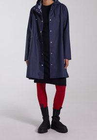 Tmavě modrý nepromokavý plášť s kapucí, drukovými knoflíky a bočními kapsami. Nosit přes černé šaty, doplněné červenými punčochami a robustními černými botami.