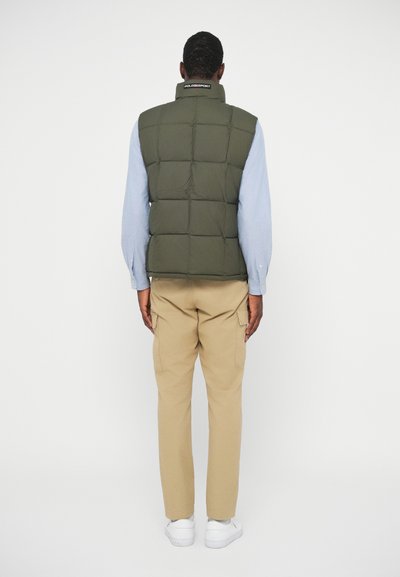 Zeleni puffer brezrokavnik s pravokotnim prešitim vzorcem, visokim ovratnikom in logotipom na vratu, v kombinaciji s svetlobradimi cargo hlačami in belimi teniskami.