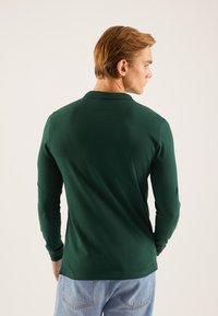 Langærmet polo i mørkegrøn, lavet af glat bomuldsstof. Har en klassisk krave og en lige kant, kombineret med lyseblå jeans.