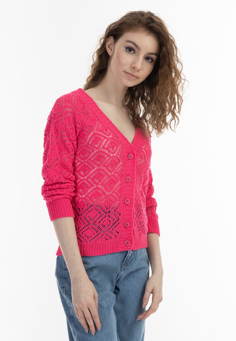 Rosa stickad cardigan med ett geometriskt mönster, V-hals, långa ärmar och fem knappar framtill. Bärs över ljusblå jeans.