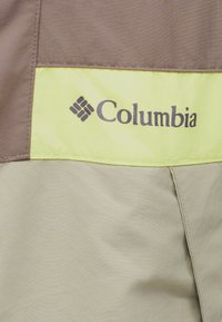 Logo Columbia sur une veste avec des panneaux en tissu marron, jaune clair et beige, avec des coutures visibles.