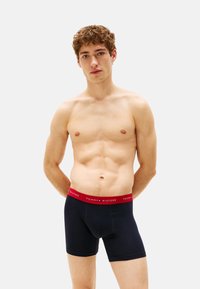 Boxers de algodón azul con una cinturilla elástica roja que presenta el logo de "TOMMY HILFIGER". Textura suave, diseño ajustado y longitud hasta medio muslo.