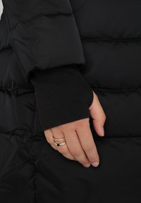 Musta toppatakki, hihassa resoranneke. Käsi, jossa on kolme sormusta, lepää takin pinnalla.