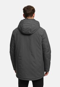 Homme portant une veste d'hiver à capuche gris foncé, debout dos à l'appareil photo sur un fond clair uni.