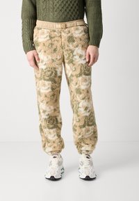 Beige un zaļo kamuflāžas rakstu polāra auduma bikses ar elastīgu jostu un pelēku jostu. Apvienotas ar baltām un pelēkām kedām.