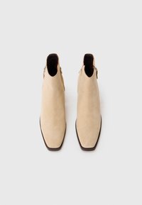 Stivaletti in suede beige alla caviglia con punta affusolata e zip laterale. Suola marrone piatta con dettagli minimi e texture liscia.