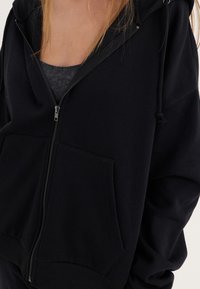 Felpa nera con zip, dotata di una ampia tasca frontale, coulisse sul cappuccio e un tessuto morbido al tatto. Il design presenta maniche a taglio ampio.
