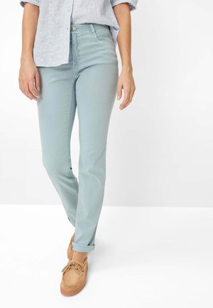 Frau trägt hellblaue Jeans mit hochgekrempelten Bündchen, hellblau gemustertes Hemd mit hochgekrempelten Ärmeln und beige Schnürschuhe, steht auf weißem Boden.