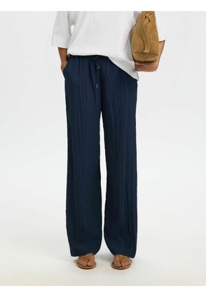 Inimene kannab lahtiseid tumesiniseid pükse, valget topi, beeži värvi sandaale ning hoiab beežist seemisnahast käekotti valgel taustal.