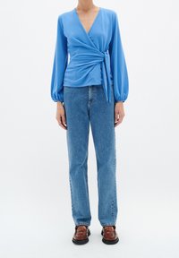 Ljusblå omslagstopp med långa, puffärmar och knytning vid midjan, tillsammans med blå jeans i rak modell och bruna flätade skor.