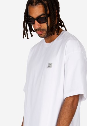 Mann mit geflochtenem Haar, der schwarze rechteckige Sonnenbrillen und ein weißes übergroßes T-Shirt mit kleinem Brustlogo trägt.