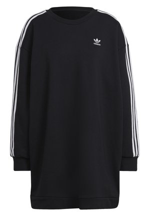 Henkilö pukeutunut mustaan Adidas-hupparimekkoon, jossa valkoiset raidat hihoissa, ja maavervoihin lenkkareihin, seisoo käsivarret ristissä ja jalat leveästi.