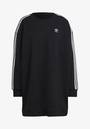 Osoba w czarnej sukience typu sweatshirt Adidas z białymi paskami na rękawach i fiołkowymi trampkami, stoi z założonymi rękami i nogami rozstawionymi na boki.