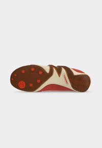 Semelle d'une basket rouge et beige avec des grips en caoutchouc marron texturés et des accents circulaires orange sur l'avant-pied et le talon.