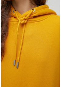 Gros plan sur un sweat à capuche jaune avec un cordon noué et des embouts métalliques, porté par une personne aux longs cheveux bruns.