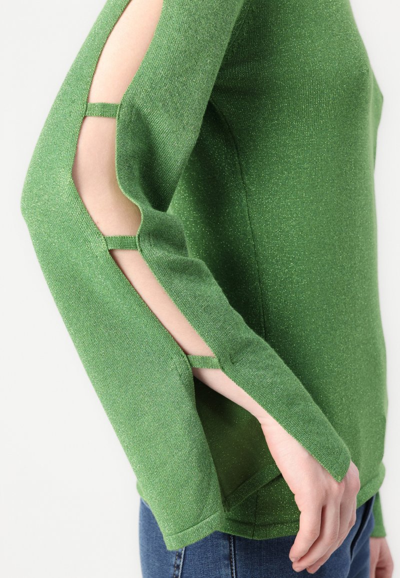 Grüner Strickpullover mit ausgeschnittenen Ärmeln und Trägern, glitzernder Textur und tailliertem Design. Sichtbare Hand, die die Kante hält.