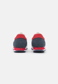 Sneaker mit einem weißen Mesh-Obermaterial, dunkelblauem Wildleder-Fersenbereich und roten Akzenten. Verfügt über eine strukturierte Sohle und kontrastierende Streifen auf der Zwischensohle.