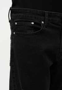 Κοντινή λήψη μαύρου τζιν με σημεία μεταλλικών πριτσίνων στην μπροστινή τσέπη και θηλιά για ζώνη, μπροστά σε απλό φόντο.