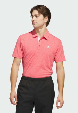 Ung mand iført en pink kortærmet polo med hvidt logo og sorte bukser, der kigger til venstre mod en ensfarvet baggrund.