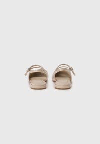 Sandale beige avec une finition en cuir brillant, dos ouvert, double brides fines avec des accents métalliques, semelle plate et texture lisse.