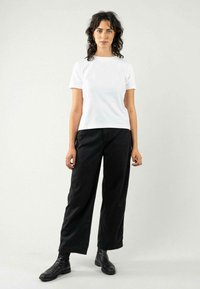 T-shirt blanc à col rond associé à un pantalon noir à taille haute et jambes larges. Le mannequin porte des bottines noires et des boucles d'oreilles en argent en forme de cercle.