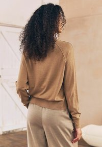 Brun strikket sweater med lange ærmer og ribbet kant, parret med lysegrå skræddersyede bukser. Baggrunden præsenterer en lys væg og trægulv.