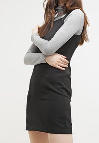 Kvinna som bär en svart ärmlös klänning med framfickor över en grå långärmad polotröja, med armarna korsade mot en vit bakgrund.