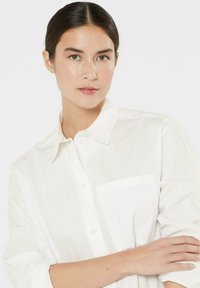 White button-up shirt featuring a single breast pocket, made of smooth fabric with a classic collar and rolled sleeves.