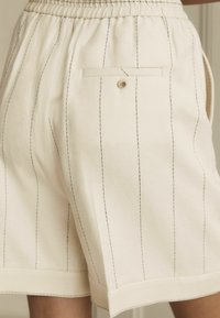 Hvide skræddersyede shorts med sorte lodrette striber, elastisk talje, sidelommer, en baglomme med knaplukning, glat tekstur.