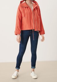 Korallenfarbene Windjacke mit Kapuze, zwei Fronttaschen und verstellbarem Bund. Kombiniert mit dunklen Jeans und weißen Sneakers.