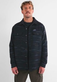 Hombre con cabello rizado y bigote, vistiendo una camisa de botones oscura con un patrón y bolsillos en el pecho, y pantalones cargo oliva, de pie frente a un fondo simple.