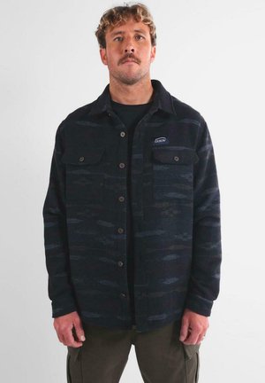 Hombre con cabello rizado y bigote, vistiendo una camisa de botones oscura con un patrón y bolsillos en el pecho, y pantalones cargo oliva, de pie frente a un fondo simple.