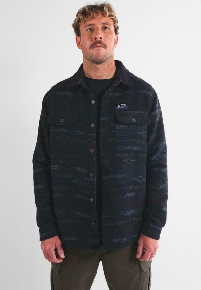 Hombre con cabello rizado y bigote, vistiendo una camisa de botones oscura con un patrón y bolsillos en el pecho, y pantalones cargo oliva, de pie frente a un fondo simple.