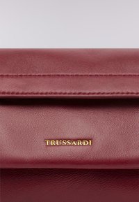 Burgundská kožená kabelka s hladkým povrchem, zlatým logem "TRUSSARDI" a designem se sklápěním s jemným prošíváním.