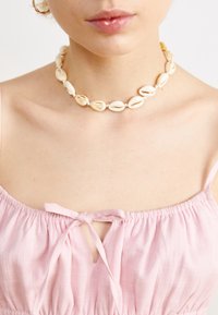 Collier ras du cou composé de coquillages blancs enfilés ensemble ; présente des surfaces lisses et des motifs naturels. Porté au-dessus d'un haut en tissu rose.