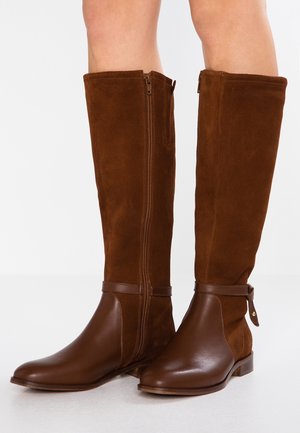 Bottes marron hautes jusqu'au genou combinant un pied en cuir lisse et une tige en daim avec des fermetures éclair latérales et des brides à la cheville sur fond blanc.