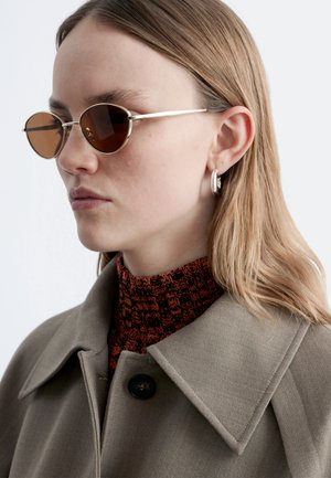 Kobieta o jasnobrązowych włosach, nosząca okrągłe okulary przeciwsłoneczne z brązowymi szkłami, srebrną kolczyk kołowy, beżowy płaszcz oraz czerwony sweter z golfem w wzory.