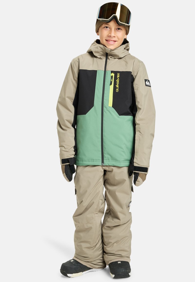 Detský lyžiarsky outfit v béžovej, zelenej a čiernej farbe. Zahŕňa bundu s kapucňou, vreckami na zips a nastaviteľnými manžetami, doplnenú o zodpovedajúce nohavice.