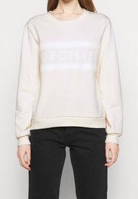 Krämfärgad sweatshirt med ribbad krage och ärmslut; har ett vitt horisontellt band med texten "CECILIE." Mjuk textil, avslappnad passform.