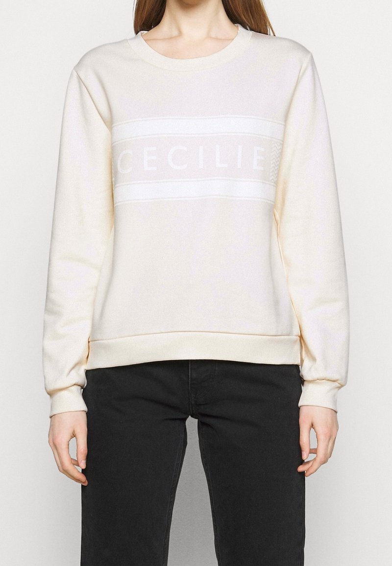 Krämfärgad sweatshirt med ribbad krage och ärmslut; har ett vitt horisontellt band med texten "CECILIE." Mjuk textil, avslappnad passform.