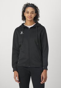 Joven con cabello rizado que lleva una sudadera negra con cremallera y pantalones negros a juego, de pie frente a un fondo liso y claro.