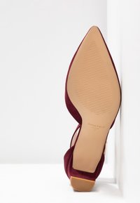 Burgundy mockask höghälssko med spetsig tå, med en texturerad sula och ankelsnodd. Varumärke synligt på sulan.