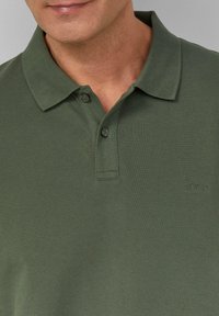 Donkergroene poloshirt van een gestructureerde stof. Heeft een klassieke kraag, twee knopen bij de hals en een klein logo op de borst.