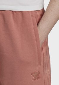 Rosa sweatpants gjorda av mjukt tyg, med sidofickor, ribbad midjesträng och en guld Adidas-logga på vänster ben.