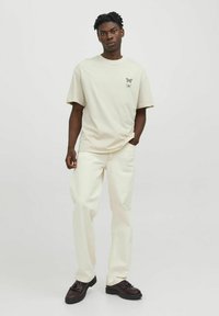 Cream-colored cotton t-shirt with a butterfly print on the chest, paired with off-white wide-leg pants and dark shoes with tassels.